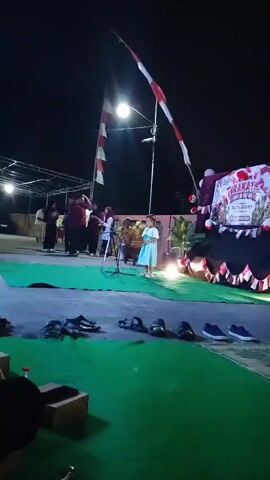 A nighttime outdoor scene shows a small girl in a light blue dress standing at a microphone on a raised platform covered in green fabric. Behind her, a larger group of people, some in dark clothing, stand near a tent-like structure decorated with red and white banners and balloons. The Indonesian flag, represented by red and white stripes on poles, is visible in the background. The ground in front of the platform has several pairs of shoes and sandals laid out. The lighting is dim and primarily artificial, with spotlights illuminating the performance area.