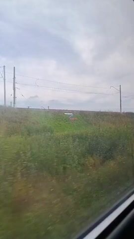 The video shows a view from a train window looking out at a grassy embankment with a cloudy sky above. There are electrical poles and wires for the train tracks running along the top of the embankment. In the distance, a faint outline of mountains can be seen against the sky. The grass on the embankment is a mix of green and brown, with some patches of yellow flowers. The train is moving, causing a slight blur in the foreground.