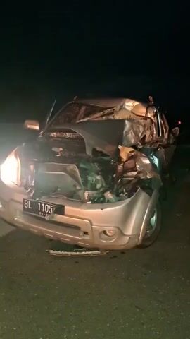 The video shows a severely damaged silver SUV at night, likely on a road. The car's front end is crumpled and appears to have sustained a major impact, with the hood open and engine components exposed. The windshield is also damaged. The license plate, which is partially visible, reads "BL 1105". A person in a blue shirt is seen standing near the front of the car, illuminated by the headlights. The scene is dark, suggesting it's nighttime, and there are no indications of weather conditions. The overall mood is somber due to the evident destruction of the vehicle. The location context indicates this event occurred in Baksukon, Indonesia.