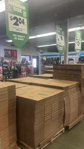 The media file depicts an indoor scene, likely a retail store. Piles of stacked cardboard boxes, prominently branded with "U-Haul" and advertising different box sizes like "MEDIUM BOX" and "LARGE BOX," dominate the foreground and midground. Green banners with pricing information, such as "$2.04" and "$8," are hanging from the ceiling. In the background, shelves stocked with various items, possibly moving supplies or home goods, are visible. The lighting suggests it is daytime, and the polished concrete floor reflects the overhead lights. The overall atmosphere appears to be one of commercial activity and preparation for moving or storage. Some text visible on the banners includes pricing and promotional messages like "WON'T BEAT!" and "BUY-IN-BULK GUARANTEE." The final frames of the media show a close-up of a person's hand making a gesture.