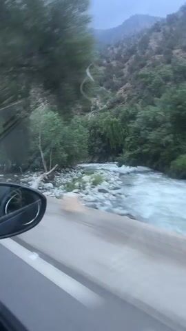 The video, shot from the perspective of a moving vehicle, captures a scenic drive along a rural road in Asgaour, Morocco. To the left, the car's side mirror is visible, reflecting a blurred view of the interior. The landscape outside the window features a lush, green environment with numerous trees and mountainous terrain in the background.

A river or stream with flowing water and rocky banks runs parallel to the road on the right side. As the vehicle moves, various objects and structures come into view: a blue metal fence or gate, followed by a small, rustic building with a white tarp roof. Further along, another light brown building is seen, with a red plastic chair placed outside it. A red flag, possibly the Moroccan flag, flies from a pole near this building. White and red striped posts are also visible along the roadside, likely marking the edge of the road. The overall impression is one of a journey through a natural, untamed landscape with intermittent signs of human presence.