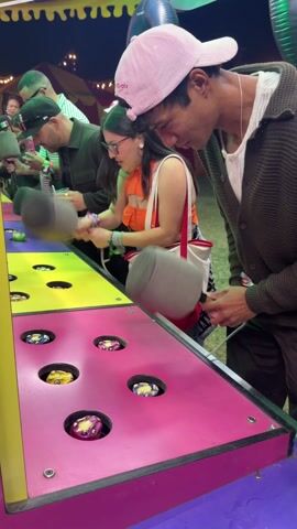 A medium shot depicts two young adults, a man in a pink baseball cap and a woman in a bright orange top and glasses, intensely playing a Whac-A-Mole game. They stand side-by-side, each wielding a gray mallet, rhythmically striking at the colorful moles popping up from holes on the game's surface. The game table is divided into yellow and pink sections, contrasting vividly with the dark gray of the mallets. Behind them, other people are engaged in the same game, including a man in a black cap and a green shirt.  A striped tent, balloons, and various carnival elements are visible in the background, indicating an outdoor fair or festival setting. The focus is on the intensity of the two individuals as they concentrate on hitting the targets in this classic arcade game.
