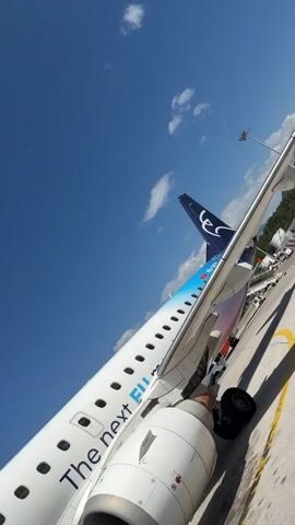 The video begins with a close-up shot of the tail of an airplane with the logo "lec" and "the next" written on the side of the fuselage. The camera pans to the side of the plane, revealing the airline's name, "AIR MONTE NEGRO," and the text "28 by 28." The scene then shifts to a group of passengers boarding the aircraft via a mobile staircase. The passengers are seen from behind, some carrying luggage. One person is wearing a white hat, another a grey tracksuit, and a third has a straw bag. A flight attendant in a yellow vest is visible in the background. The video concludes with a shot from the perspective of someone walking on the tarmac, their legs and feet in white pants and sneakers, with luggage visible in the foreground. The environment appears to be an airport tarmac on a clear, sunny day.