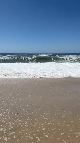 A wide shot of a sandy beach with ocean waves rolling in. The sky is clear blue. The waves are moderate, with white foam cresting as they break and wash onto the shore. The sand is light brown with small pebbles and shells visible. Bubbles from the receding waves are scattered on the wet sand. The video is shot from a low angle, looking out towards the ocean.