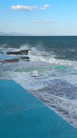A choppy ocean with waves crashing against a concrete breakwater. The water is a deep blue-green, and the sky is a clear blue with a few wispy clouds. The breakwater appears to be made up of several tiered levels of concrete, some of which are painted in vibrant colors like turquoise and purple. The waves are breaking in white foam as they hit the structure, with some spray rising into the air. The overall scene conveys a sense of raw, natural power meeting man-made construction.