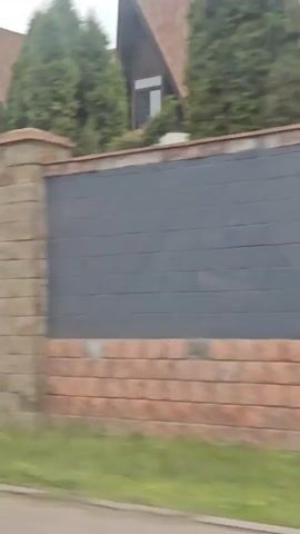 A video pans across a property with a tall fence made of brick and dark grey panels. On top of the fence, evergreen trees are visible, with the steep, brown roof of a house peeking out behind them. The house has triangular gables and windows. A decorative black metal gate with ornate scrollwork is seen on the right side of the fence. The scene appears to be outdoors during the day, with an overcast sky.