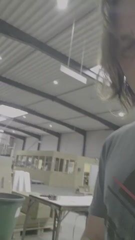 The video shows a close-up of a person's face, who is wearing a gray t-shirt. The background appears to be a warehouse or industrial space, with a metal ceiling, lights, and some structures resembling offices or workstations. The person is looking directly at the camera and their expression is neutral. The camera movement is minimal, primarily focusing on the person's face.