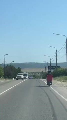 A white van is traveling on a highway. A person on a red scooter is in front of the van. There are other vehicles on the road and trees on the side of the road. The sky is clear and blue.