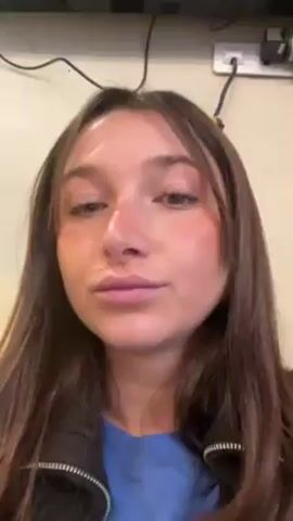 A young woman with long brown hair and fair skin, wearing a blue top and a black jacket, is the primary subject of this close-up video. Her face is slightly flushed, particularly on her cheeks. She opens her mouth, then looks upward before holding a yellow pill near her mouth. The background is a plain wall with an electrical outlet and some wires.