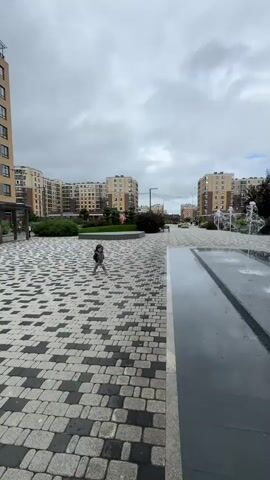 A young child runs across a paved area in front of apartment buildings. The child is wearing a grey vest over a blue and white shirt and patterned pants. The ground is made of cobblestones with a repeating pattern of light and dark grey squares. In the background, there are modern apartment buildings with many windows. To the right, there is a water feature with jets of water spraying upwards, and a dark, wet surface. The sky is overcast with grey clouds. The child is holding a small black object in their right hand. The child appears to be walking towards the camera at the end of the clip.