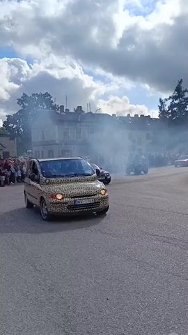 A leopard-print Fiat Multipla is performing a drift, emitting a significant amount of tire smoke. A crowd of spectators is visible in the background, lining a street in front of buildings. The sky is cloudy.