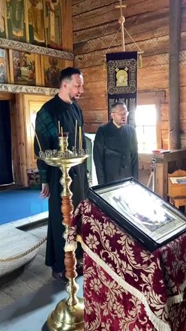 Two men in dark robes stand inside a wooden church. The man on the left is facing the camera and speaking, with a lit candelabra in front of him. The man on the right is facing the icon on the right, singing or chanting. The icon is on a stand draped with a patterned cloth. Behind them are icons on the walls and a banner hanging from the ceiling. The lighting is natural, coming from a window on the right.