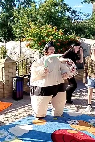 This media file captures a playful outdoor scene featuring two individuals in inflatable sumo wrestler costumes. One person, wearing a cream-colored suit with a black belt, is holding a green balloon sword and looking towards the camera with a smile. The other person, also in a sumo costume, is facing away from the camera. They appear to be engaged in a friendly sparring match or a game.

The setting is a backyard with a brick fence on the left and lush green trees and bushes in the background. Sunlight suggests it's daytime, and the clear sky indicates fair weather. A bicycle is visible leaning against the fence. The ground is covered with a colorful mat, possibly for children's play, with cartoon-like figures. A black and blue object, possibly a cooler or storage bin, is also present near the fence.

The overall mood appears to be lighthearted and joyful, with the participants seemingly enjoying their playful activity. The inflatable costumes, balloon swords, and the casual outdoor setting all contribute to a sense of fun and recreation. No visible text is present in the media file.