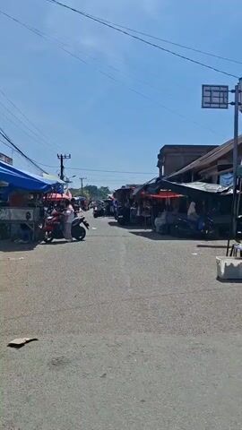 The video shows a street market on a sunny day. The street is paved and lined with stalls on both sides. The stalls are covered with tarps and umbrellas in various colors, including blue, red, and brown. Various goods are displayed, such as clothing and food items. Several motorcycles are parked along the sides of the street and some are moving. People are seen walking around and interacting with the vendors. Some people are wearing traditional clothing. The sky is clear and blue. The overall atmosphere is that of a busy, outdoor marketplace.