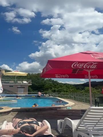 This is a video of a swimming pool area on a sunny day. In the foreground, there are lounge chairs and umbrellas. One umbrella is white with Bulgarian text, and another is red with a Coca-Cola logo. A person in a black bikini is lying on a lounge chair, facing away from the camera. In the background, there is a blue swimming pool with several people in it, some wearing inflatable rings. There is a building with a balcony and pink umbrellas on the left side of the frame, and lush green trees and vegetation covering a hillside behind the pool. The sky is blue with white clouds.