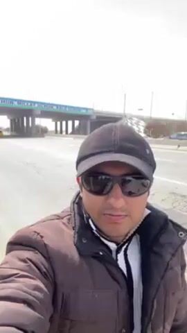 A man in a brown jacket, black cap, and sunglasses takes a selfie on a road. Behind him is a bridge with the words "Vatan uchun, Millat uchun, Xalo uchun" printed on it. Cars drive on the road under the bridge. The sky is bright and overcast.