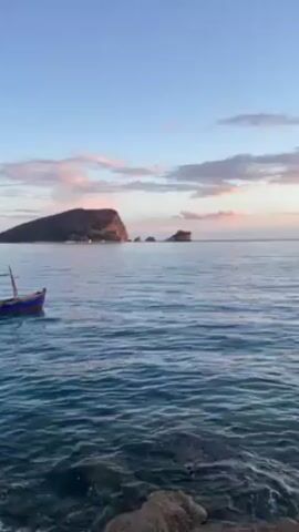 The video shows a small motorboat cruising through calm blue water towards the horizon. In the distance, a large rocky island stands tall, accompanied by a few smaller rock formations. The sky above is a blend of light blue and pink hues, indicative of either sunrise or sunset. A person is visible in the boat. The foreground consists of rocks along the shoreline.