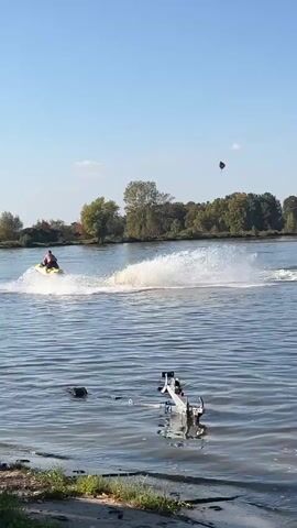 A jet ski is ridden by a person wearing a red shirt on a wide body of water under a clear blue sky. The jet ski is leaving a white wake as it moves from left to right across the frame. In the foreground, on the right side of the water, a white mechanical device with wheels is partially submerged. In the background, a treeline with green and yellow foliage borders the water. A person's silhouette can be seen on the left bank of the water in the foreground.
