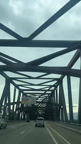The video shows a view from a car driving across a bridge. The bridge is a large metal truss bridge with a complex structure of intersecting beams. The sky is overcast with clouds. Several cars are visible on the road ahead, including a white pickup truck in front of the camera car. There is a toll sign visible on the left side of the road, indicating that the toll for cars is $1.65. The road appears to be a multi-lane highway. The camera is pointing forward and slightly upwards, capturing the bridge structure and the road ahead.