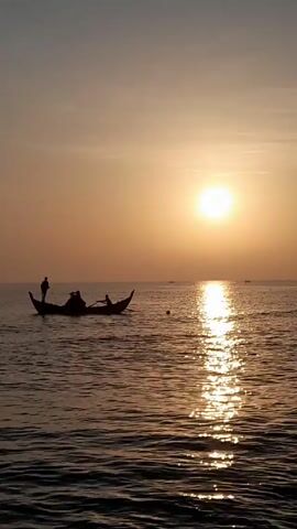 The video shows a small boat with three silhouetted figures on board, moving across the ocean at sunset. The sun is a bright, hazy circle in the sky, casting a long, shimmering reflection on the water. The sea is calm with gentle ripples. The sky is a gradient of light orange and grey. There are a couple of distant boats in the background.
