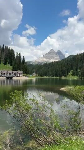 The video captures a serene landscape with a clear blue sky dotted with fluffy white clouds. In the foreground, a calm lake reflects the sky and surrounding scenery. On the left side of the lake, there is a large building with a distinctive roof structure, possibly a hotel or restaurant, nestled amongst green trees. Across the lake, a dense forest covers the rolling hills, leading up to a majestic mountain peak in the distance. The mountain appears rugged with visible rock formations. The overall environment is natural and picturesque, suggesting a tranquil setting, possibly in a mountainous region during daytime. There are no people or animals visible in the frame.