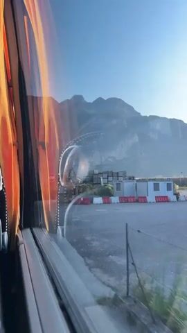 A video begins from the perspective of someone inside a vehicle, looking out the window. The window frame and an orange curtain are visible on the left side of the screen. Outside, a clear blue sky stretches above rugged mountains in the background. In the middle ground, there are industrial elements like a truck with a crane, piles of wooden planks, and a red and white stop sign. A road sign that reads "MEZZOLOMBARDO" is also visible. The landscape transitions to tall green grass and vegetation in the foreground as the vehicle moves. The footage shows a brief moment of travel, passing by signs of infrastructure and a town name in a mountainous region.