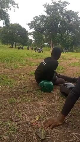 A group of uniformed individuals, likely police officers, are gathered in an open grassy field with trees in the background. In the foreground, two individuals are seated on the ground. One individual, facing away from the camera, is wearing a black shirt with white lettering that reads "POLICE." This person is sitting on a green helmet. Another individual's arm and shoulder are visible in the lower right corner of the frame. Several other uniformed individuals are dispersed throughout the field, some appearing to be in formation or engaged in some activity. The sky is overcast.