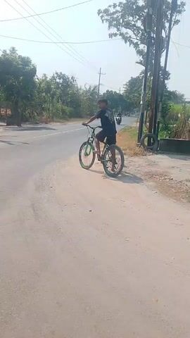 A person on a bicycle turns a corner on a dirt road next to a paved road. There are trees and power lines in the background. Another person on a scooter passes by. The camera then shifts to a first-person view from the scooter, showing the dashboard.