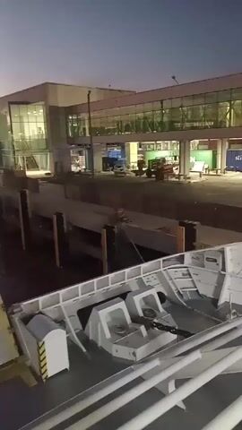 The video shows a ferry at a port or dock at dusk or night. The camera pans across the top deck of the ferry, showing empty seating, windows revealing passengers inside, and railings. The camera focuses on the view from the ferry, revealing a nearby building and another ferry labeled “Baquebus”. The sky is a mix of dark blue and pink hues. The water reflects the lights of the surrounding structures, indicating calm conditions.
