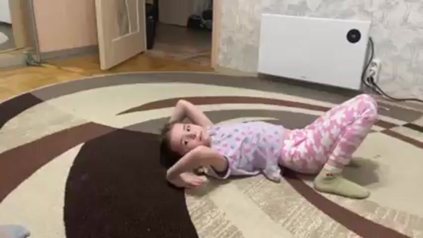 A young girl is performing a gymnastic backbend on a patterned, circular rug. She wears a light-colored pajama set with a flower pattern and mismatched socks. Her hair is braided, and she looks upwards as she arches her back, supporting herself with her hands and feet. Behind her is a white radiator mounted on a wall with patterned wallpaper. A partially open door reveals a glimpse into another room. The lighting is indoor and fairly dim.