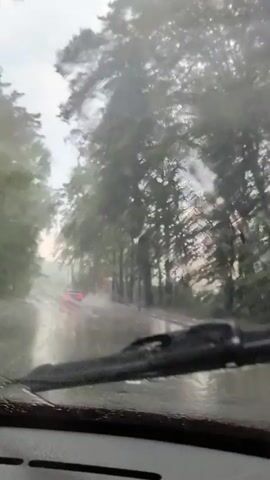 The video captures a view from inside a car driving on a wet road during heavy rainfall. The scene is set on a tree-lined street, with lush green foliage on either side of the road. The road itself appears to be flooded in places, with water splashing up as the car moves. A red car is visible in the distance, its taillights illuminated. The sky is overcast, indicating a cloudy and possibly stormy day. The time of day is difficult to determine precisely, but the overcast conditions suggest it could be daytime. There is no visible text in the video. The overall mood conveyed is one of a challenging drive due to adverse weather conditions.