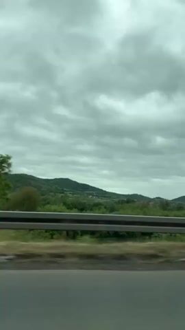 The video shows a view from inside a moving vehicle, likely a car, driving alongside a highway. A gray guardrail runs along the edge of the road. Beyond the guardrail, a landscape unfolds, featuring green hills, trees, and vegetation under an overcast sky filled with gray clouds. The road surface is visible in the foreground. The scene is static in terms of the camera position, with the movement implied by the passing scenery.