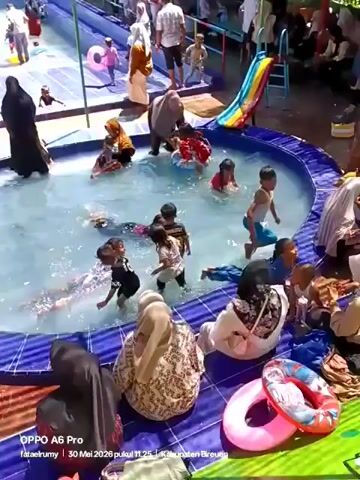 Here's a description of the video:

The video shows a crowded swimming pool area. There are two pools visible, one larger and one smaller, both filled with people of various ages, including many children playing and swimming. Some people are standing or walking around the pool. Some are wearing hijabs. In the foreground, people are sitting near the edge of the smaller pool. One man is holding a baby while talking to someone else. The pool area appears to be outdoors and is constructed with blue and white tiles. A small slide is visible in the background by the larger pool. There's a lot of splashing and general activity in the water.