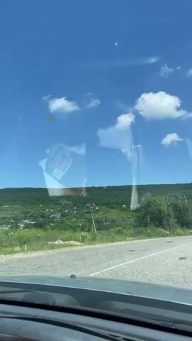 The video shows a view from inside a vehicle, looking out through the windshield. The vehicle is driving on a paved road with a white line down the center. To the left of the road, there is a grassy area and then a dense green forest covering rolling hills. The sky is a clear bright blue with scattered white cumulus clouds. Sunlight reflects off the windshield, obscuring some details. In the distance, on the right side of the road, another white vehicle is visible. The overall scene depicts a scenic drive through a natural landscape on a sunny day.