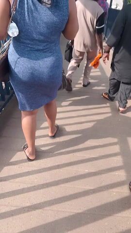 The video shows a low-angle perspective of people walking on a paved surface, likely a sidewalk, with a metal railing to the left and shadows cast by a fence or structure to the right. A woman in a blue dress and flip-flops is visible, carrying a purse. Another person, wearing striped pants, is seen from behind, holding an orange object. Several black plastic bags are on the ground. The camera is tilted, giving a sideways view of the scene.