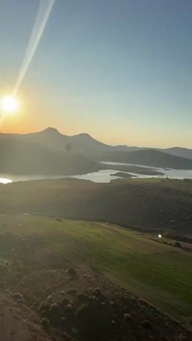 The video captures a breathtaking hot air balloon flight over the scenic landscape of Yeşilhisar, Turkey, during sunrise. The early morning light bathes the entire scene in a soft, golden glow, with a visible sun flare emanating from the left side of the frame.

The panoramic view showcases rolling hills, distant mountains, and a serene body of water, possibly a lake or reservoir, nestled within the terrain. Below, a patchwork of agricultural fields, some vibrantly green and others with earthy tones, stretches across the foreground.

As the camera slowly pans from left to right, multiple hot air balloons appear, gracefully ascending into the clear blue sky. One prominent hot air balloon, distinguished by its vibrant yellow and red colors, becomes a focal point, rising above the rugged hilltops. The serene activity of the balloons dotting the expansive sky creates a captivating and tranquil atmosphere.

In the background, the silhouette of the mountains adds depth to the landscape. A person can be heard speaking in Russian, remarking "So beautiful," further emphasizing the awe-inspiring nature of the experience. The video perfectly encapsulates the beauty and tranquility of a hot air balloon journey at dawn.