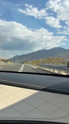 The video shows a view from the inside of a car looking out the windshield. The car appears to be on a highway, with multiple lanes of asphalt stretching into the distance. White lines mark the lanes, and a guardrail runs along the right side of the road. In the background, there are mountains covered in trees and foliage, with a bright blue sky filled with scattered white clouds overhead. Streetlights are visible along the side of the highway. The dashboard of the car is visible in the foreground, with a textured, beige material and a dark grey area. The car is in motion, indicated by the blurred scenery and the dynamic perspective. There are no people visible in the video.