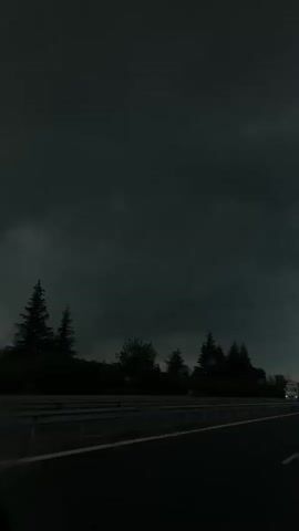 The video shows a view from a moving vehicle on a highway. The sky is dominated by dark, ominous storm clouds. The highway is black asphalt with white lane markings. A guardrail runs along the side of the road, separating the highway from a line of trees. Other vehicles are visible in the distance. Raindrops are visible on the windshield.