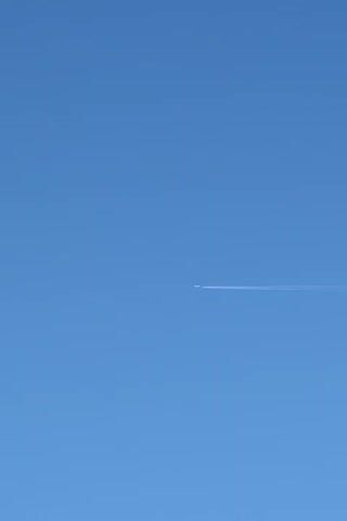 A clear, blue sky with a single airplane leaving a contrail across the frame. The airplane is small and appears to be in the distance. The contrail is white and stretches horizontally across the image. The video is static, with no visible movement except for the faint dissipation of the contrail.