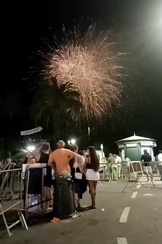 This is a night-time video of a crowd watching a fireworks display. The foreground shows a metal barrier, behind which are several people. The largest group consists of four individuals: a person in an orange shirt facing away from the camera, a person in a black tank top and shorts, a person in a white tank top and white skirt, and a person with long dark hair. To the left of this group, a person in a dark top and shorts can be seen. Further left, a few more people are visible behind the barrier. In the middle ground, a palm tree is silhouetted against the night sky, with a banner hanging from it. Beyond the crowd, a small, ornate white kiosk with green trim is visible, as are more people and barriers. The background is dark, with the bright bursts of fireworks illuminating the sky. The fireworks are a mix of large, fanned-out explosions and smaller, more clustered bursts, appearing in shades of gold, white, and red. The ground is paved and appears to be a street or promenade, with white lines painted on it. Some litter, including a plastic bottle and a crumpled piece of paper, is visible on the ground near the group in the foreground. The overall impression is of a lively outdoor event with a festive atmosphere.