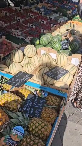 The video shows a fruit stand at an outdoor market. There are various fruits displayed, including strawberries, peaches, apricots, watermelons, cantaloupes, pineapples, bananas, kiwis, avocados, and grapes. The fruits are arranged in boxes and crates. Some of the fruits have price tags. A person in a patterned dress and a black jacket is partially visible on the right side of the frame, reaching towards the fruit. The ground is paved with cobblestones, and there are metal frames and carts visible in the background. The scene is well-lit, suggesting it is daytime.