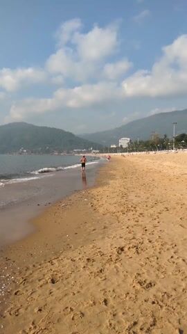 The video shows a beach scene on a sunny day. The beach is sandy, and there are gentle waves lapping the shore.  In the foreground, the sand is marked with footprints. A man wearing shorts is jogging along the water's edge, away from the camera. In the background, there are green hills and buildings along the shoreline. Other people can be seen scattered along the beach, including another person jogging in the same direction as the man. The sky is blue with scattered clouds.