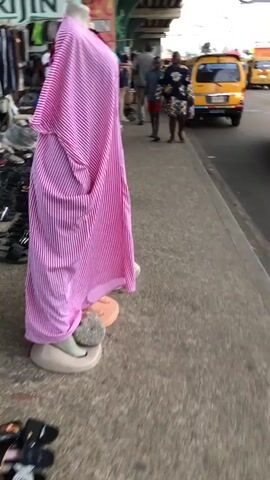 The video captures a bustling street market scene, possibly in a developing country, with a focus on clothing and shoe stalls lining a sidewalk. A mannequin draped in a pink striped fabric stands prominently in the foreground. Beyond it, various clothing items, including shirts, dresses, and pants, are displayed on the ground and on stands. A large assortment of shoes, ranging from sneakers to sandals, is neatly arranged along the edge of the sidewalk. Several people are browsing the merchandise, including two individuals walking away from the camera, one wearing a patterned dress and the other in a red shirt and shorts. A yellow taxi is visible on the road in the background, suggesting a busy urban environment. The overall impression is one of vibrant commerce and everyday life unfolding in a public space.