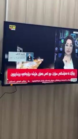 A flat-screen television displays a news broadcast from NRT News. The broadcast shows a female news anchor on the right side of the screen, wearing a black blazer and glasses. A smaller box on the left side of the screen shows a man in a suit. The screen displays text in an unknown language, with a red banner at the bottom. The television is mounted on a light brown, textured wall. The environment appears to be indoors, possibly a home.