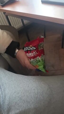 The video depicts a classroom scene. The video starts with a person in gray sweatpants holding a bag of "Beskidzkie" tzatziki flavored chips. The camera pans to show a student sitting at a desk using a phone with a water bottle placed on the desk next to her. The camera then shows a wider view of the classroom, with multiple students seated at desks, some using phones. A teacher stands in the doorway to the classroom, seemingly addressing the students. The classroom has light yellow walls, a bulletin board, and wooden desks.