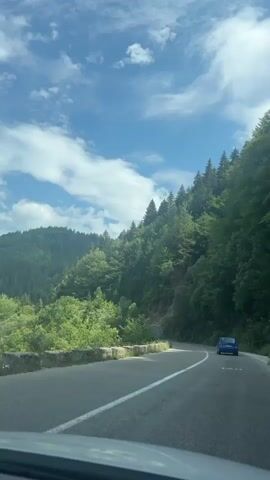 The video shows a blue car driving on a paved road that curves through a mountainous, forested area. The road has white lane markings. On the left side of the road, there is a stone wall and vegetation. On the right side, the terrain slopes upwards, covered in dense green trees. The sky is blue with scattered white clouds. The car is moving away from the camera and is in the right lane of the road. The camera appears to be inside another vehicle, looking forward. The image is well-lit, suggesting it is daytime. There are no people visible outside the vehicles. The overall scene depicts a scenic drive through a natural landscape.
