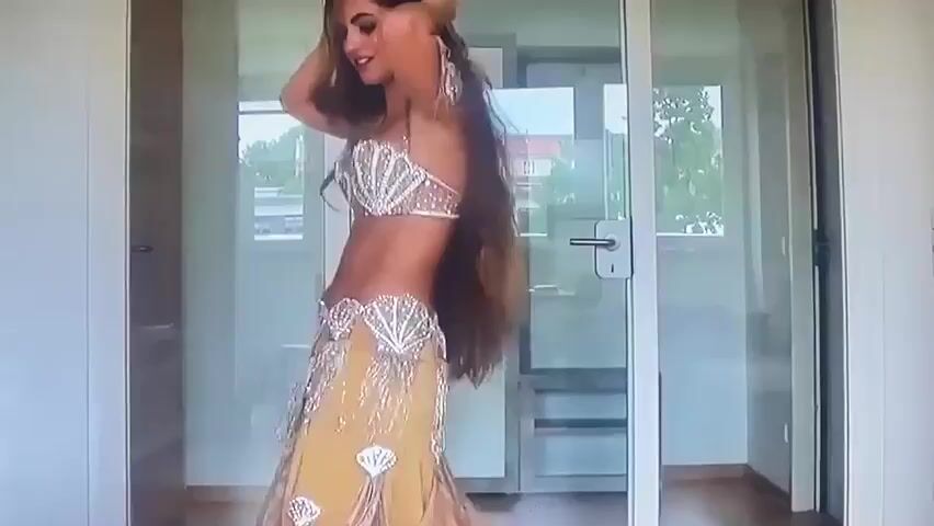 A woman with long brown hair is dancing in front of a glass door. She is wearing a sparkly gold and white belly dancing costume. The costume consists of a seashell-shaped bra, a decorated belt, and a flowing skirt. She is moving her arms and body to the music. The background shows an indoor space with wooden floors and a glimpse of an outdoor area through the glass.