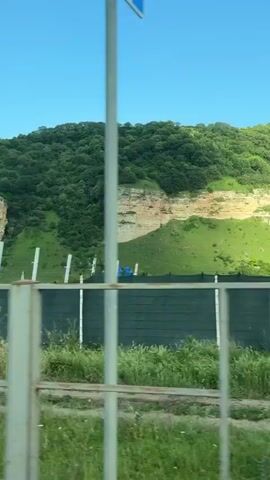 This is a vertical video clip shot from a moving vehicle. The scene captures a rural landscape with rolling green hills in the background, topped with dense, dark green trees. In the mid-ground, there are two prominent, layered rock formations that appear to be cliffs, with a gap between them. A light-colored, possibly dirt or gravel road is visible in the foreground.

A fence runs horizontally across the middle of the frame, composed of vertical white posts and dark mesh netting. Beyond the fence, there are patches of green grass and wildflowers, some of which are small and white, and a few taller purple flowers.

To the right of the frame, in the foreground, is a building with a stone base and a lighter-colored roof. Next to it is a very large, leafy green tree. A utility pole with wires is also visible near the building and tree.

The sky is a clear, bright blue, suggesting a sunny day. The lighting indicates it is likely daytime, possibly late morning or early afternoon, given the brightness and the shadows. The overall mood is peaceful and natural, showcasing a rural, possibly mountainous or hilly, environment. There is no visible text in the video.
