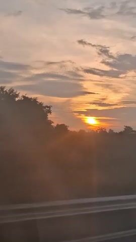 The video shows a sunrise viewed from a vehicle, likely a car, moving along a road. The sun is a bright orange disc peeking over a line of trees and foliage that forms the horizon. The sky is filled with clouds, some of which are tinged with orange and pink from the sunrise. The foreground shows the road and a barrier, suggesting the perspective is from inside the vehicle, looking out the window. The overall impression is of a peaceful and scenic drive at the beginning of the day.