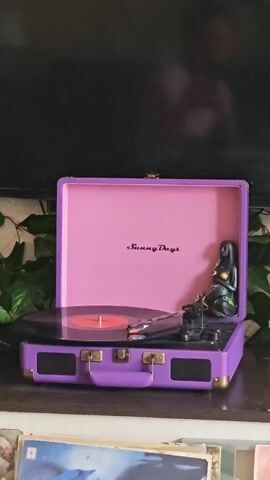 Here's a description of the video:

The video shows a purple "Sunny Days" record player with its lid open, playing a record. The record is black with a red label. A small, dark-colored figurine of a woman is positioned inside the lid of the record player. Green ivy trails around the record player's base. The record player is sitting on a wooden surface in front of a dark television screen. Several record albums are visible on the shelf below.